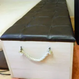 Tapicerowana pufa ze schowkiem, obita brązową skórą ekologiczną z pikowaniem, z linowym uchwytem, stojąca na dywanie.