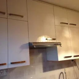 Białe szafki kuchenne z drewnianymi uchwytami, widoczny okap podszafkowy i fragment zlewu. Na ścianie płytki imitujące kamień.