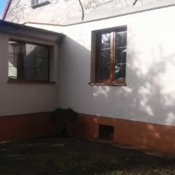 Fragment elewacji domu z brązowym cokołem, drewnianymi oknami i cieniem drzew na ścianie.