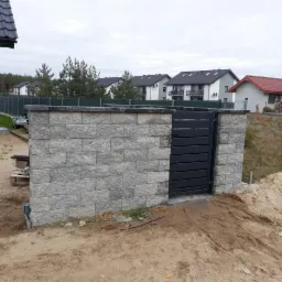 Nowo wybudowane ogrodzenie z szarej cegły z nowoczesną, poziomą bramą w kolorze antracytowym, na tle domów jednorodzinnych w jesiennej scenerii.