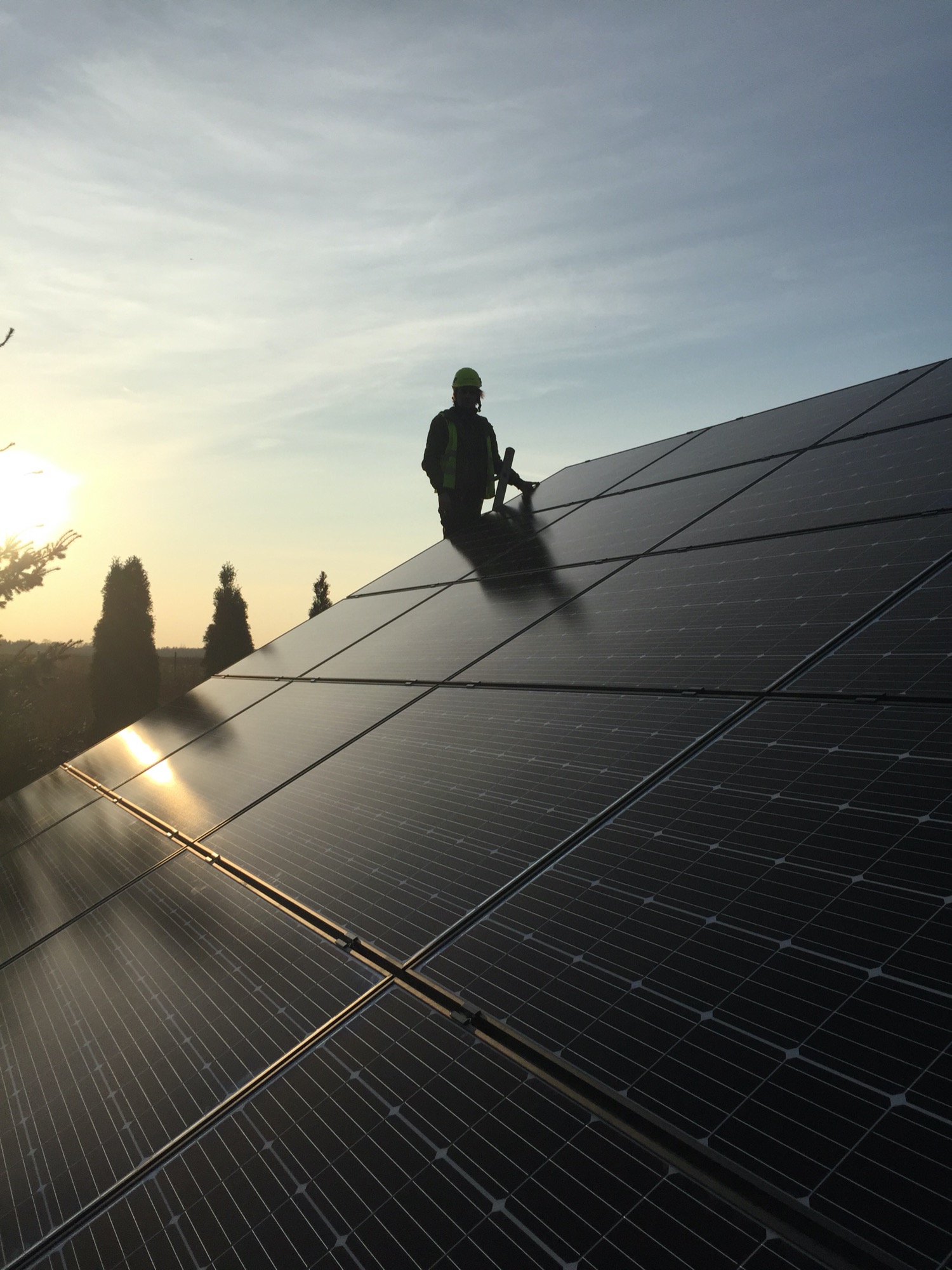 Instalator w kamizelce odblaskowej i kasku na dachu z panelami słonecznymi podczas zachodu słońca, widoczne drzewa w tle.
