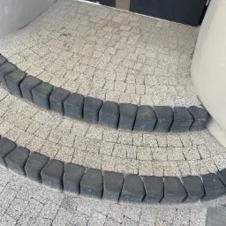 Dwa stopnie schodów wejściowych wyłożone jasną kostką brukową z ciemnoszarą obwódką, prowadzące do czarnych drzwi wejściowych.