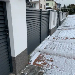 Nowoczesne, szare ogrodzenie lamelowe z bramą i białymi słupkami, wykończone dekoracyjnym tynkiem, na tle ośnieżonej kostki brukowej i zabudowy jednorodzinnej.