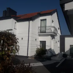 Nowoczesny dom jednorodzinny z białą elewacją, balkonem z kutą balustradą i czerwoną dachówką, widok z poziomu gruntu przez zaschnięte pnącza.