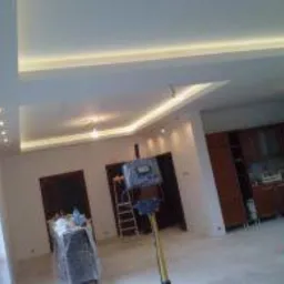 Nowoczesne wnętrze po remoncie: biały sufit z podwieszanymi elementami oświetleniowymi LED, widoczny sprzęt budowlany i drabiny, częściowo umeblowana kuchnia w tle.