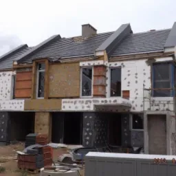 Budynek w trakcie ocieplania elewacji z widocznymi warstwami izolacji, siatką i styropianem, rusztowania przy ścianie bocznej, stosy dach&oacute;wek na ziemi.