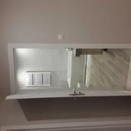 Otwarta biała łazienka widziana z korytarza, białe ściany, grzejnik drabinkowy, drewnopodobne płytki podłogowe w łazience i parkiet w korytarzu.