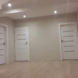 Jasne wnętrze po remoncie z trzema białymi drzwiami z poziomymi frezami, drewnianymi schodami z metalową balustradą i parkietem w odcieniach beżu.