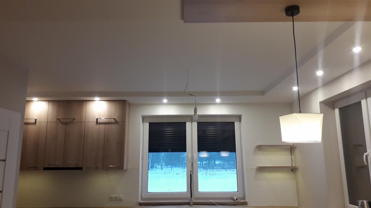 Wykończone wnętrze kuchni z drewnianymi szafkami, oświetleniem punktowym w suficie podwieszanym i nowoczesną lampą wiszącą nad oknem z roletami.