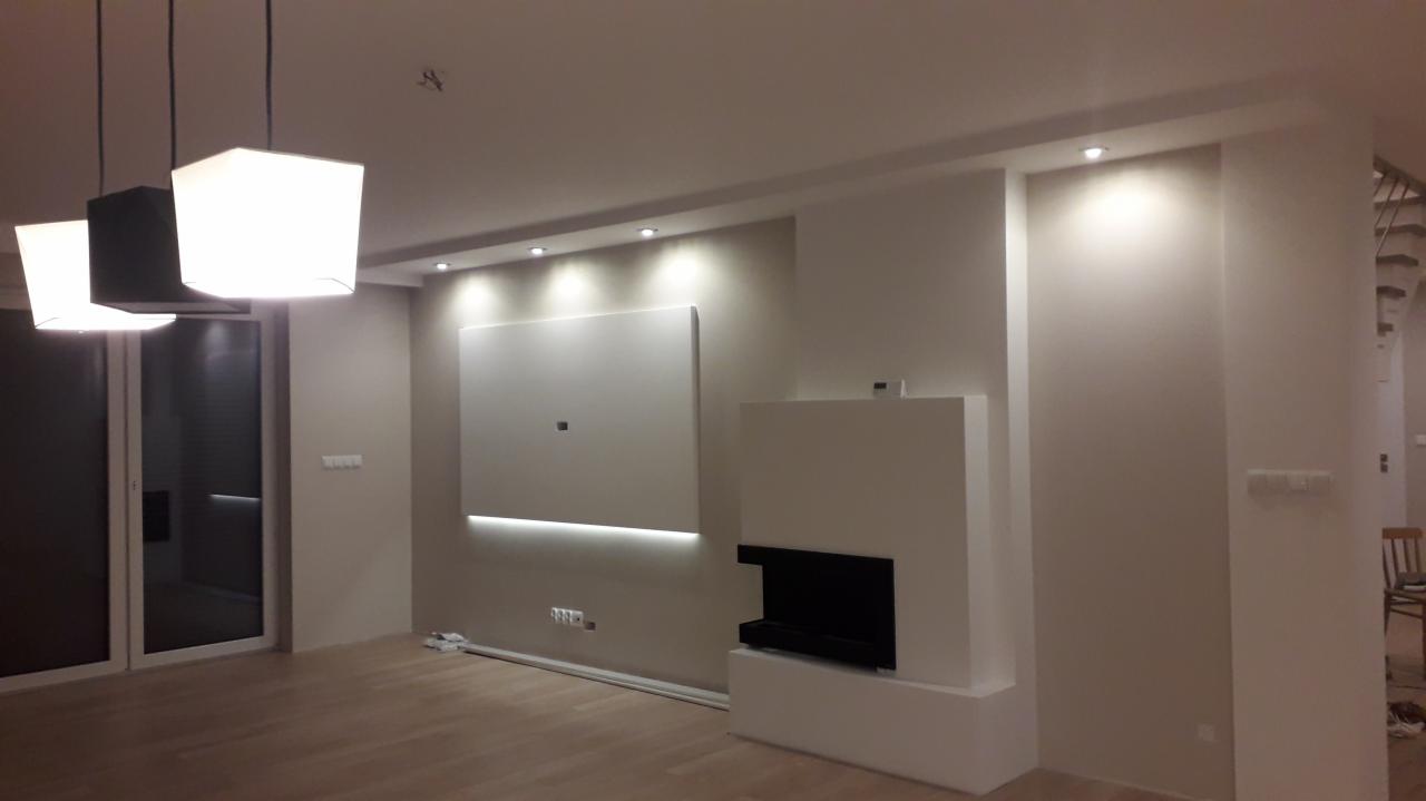 Nowoczesny salon z minimalistycznym kominkiem, centralnie umieszczonym panelem TV z subtelnym podświetleniem LED i trzema lampami wiszącymi o geometrycznych kształtach.