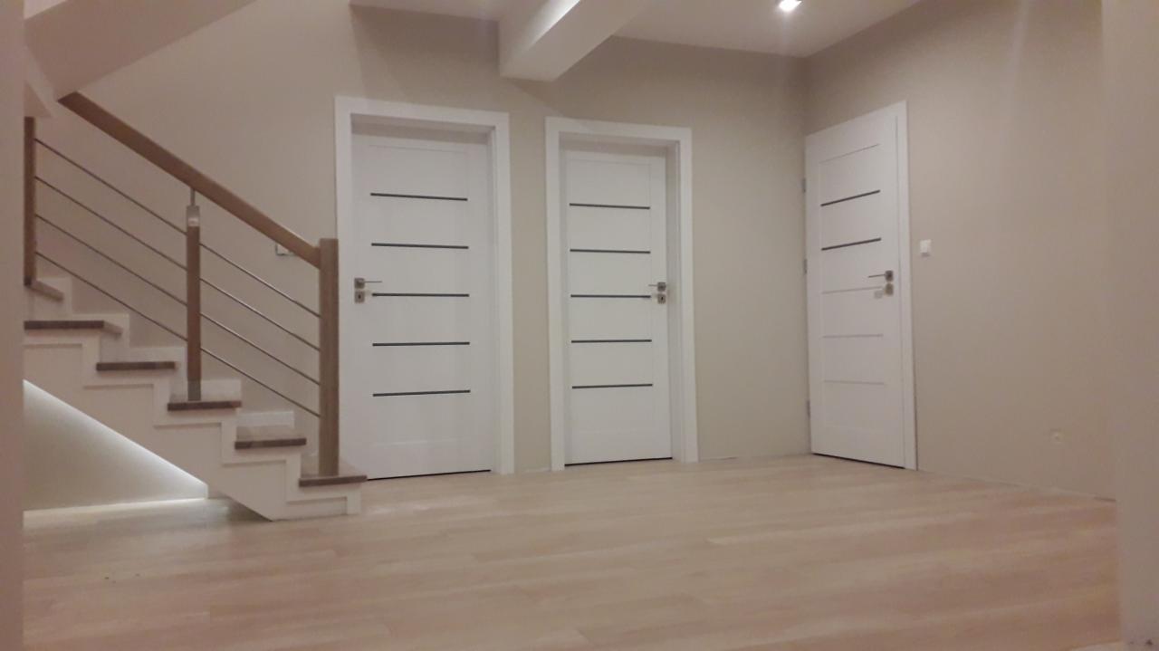 Jasne, nowocześnie wykończone wnętrze: schody z drewnianymi stopniami i metalową balustradą, trzy białe drzwi z poziomymi, czarnymi paskami, jasna drewniana podłoga.