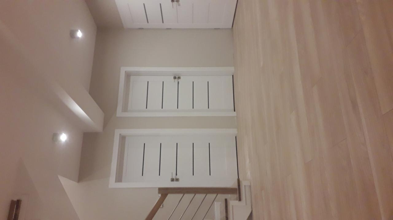 Wykończone wnętrze klatki schodowej z drewnianą podłogą, białymi drzwiami szafek w zabudowie i oświetleniem punktowym.