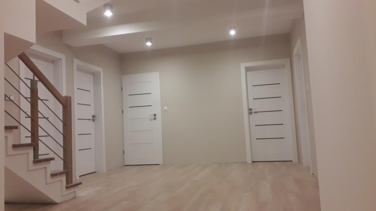 Jasne wnętrze po remoncie z trzema białymi drzwiami z poziomymi frezami, drewnianymi schodami z metalową balustradą i parkietem w odcieniach beżu.