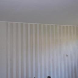 F.h.u.magbud - Ściana z tapetą w pionowe, beżowe pasy, wykończona białą listwą przysufitową, widoczne gniazdko elektryczne.