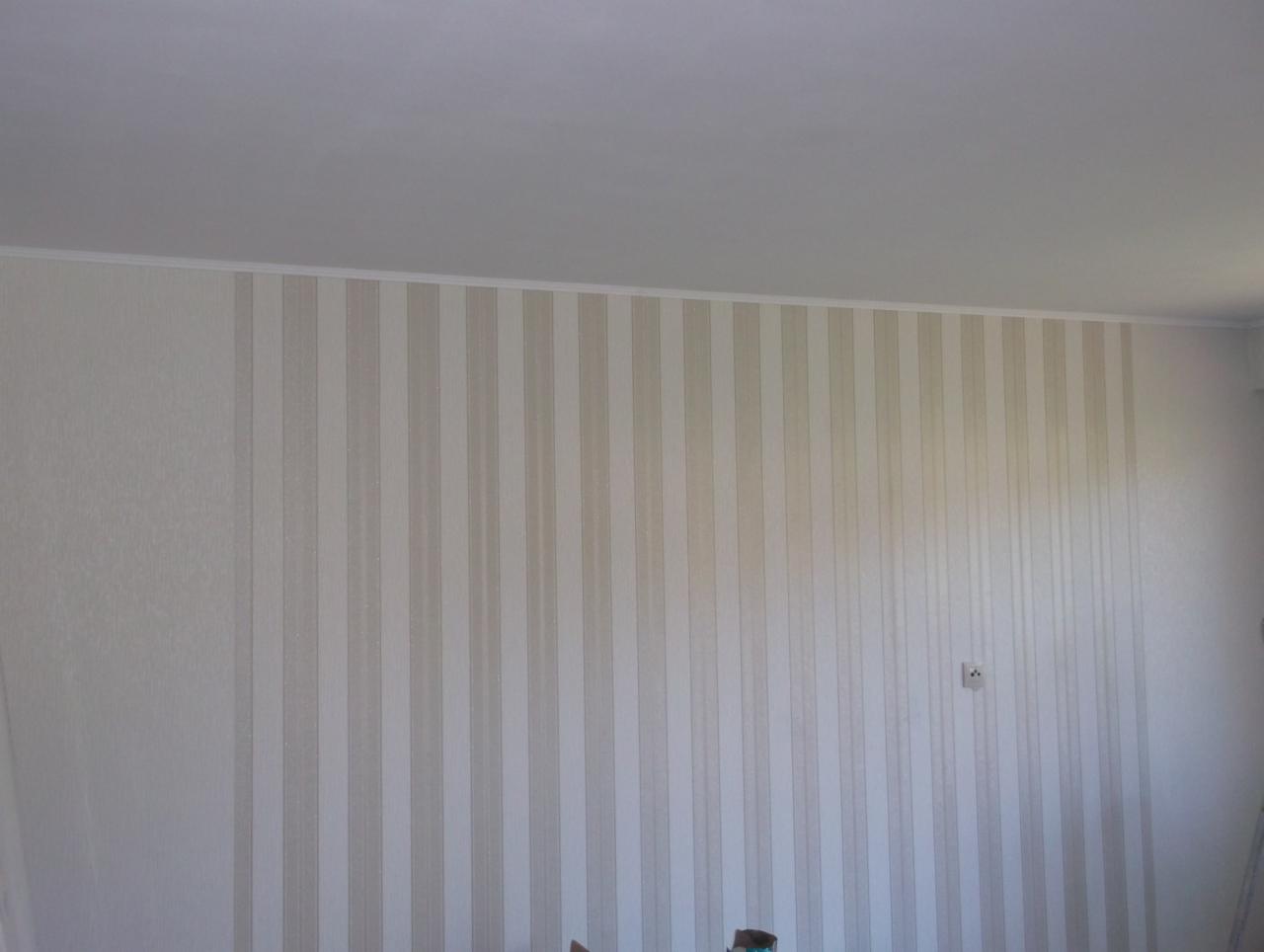 Ściana z tapetą w pionowe, beżowe pasy, wykończona białą listwą przysufitową, widoczne gniazdko elektryczne.