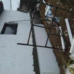 Ocieplanie budynku styropianem: widoczna ściana z przyklejonymi płytami, fragment rusztowania i resztki zaprawy w spoinach, ziemia z odrobiną śniegu.