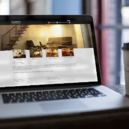 Laptop wyświetlający stronę internetową Dreamtime Apartments z widokiem na wnętrza apartamentów, obok kubek z kawą na drewnianym blacie.