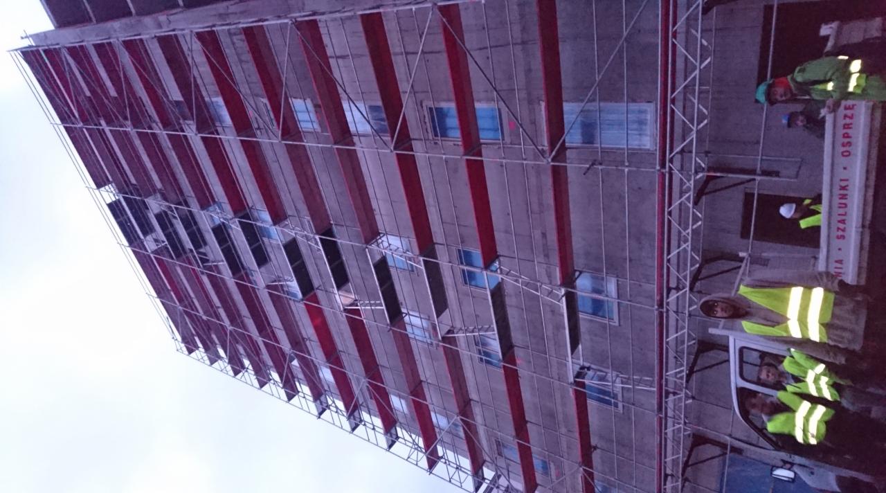 Wysoki budynek w trakcie budowy, z rusztowaniami i pracownikami w odblaskowych kamizelkach, widziany z dołu pod kątem.