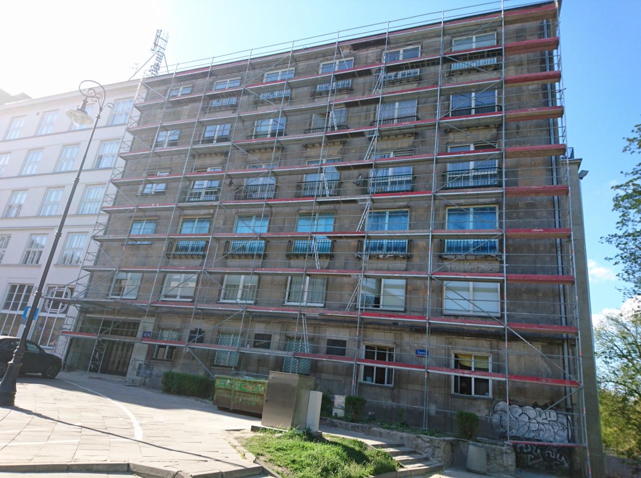Wielopiętrowy budynek w szarym kolorze, otoczony rusztowaniami, z widocznymi balkonami i oknami, sfotografowany w słoneczny dzień.