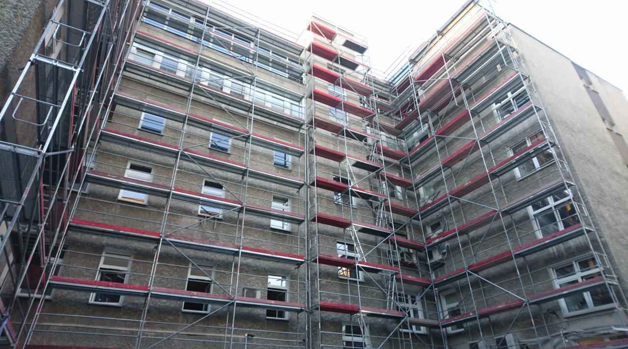 Wielopiętrowy budynek mieszkalny w trakcie remontu, osłonięty rusztowaniami z czerwonymi poprzeczkami, widok z dołu.