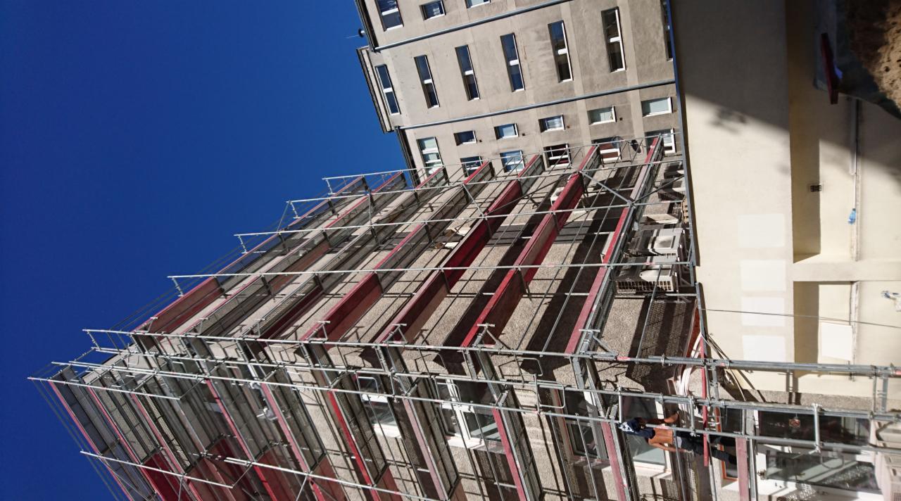 Budynek w rusztowaniach z widocznym fragmentem drugiego budynku i intensywnie niebieskim niebem, ujęcie z dołu.