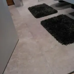 Beżowe płytki podłogowe ułożone w łazience, widoczne dwa czarne, puszyste dywaniki, fragment kabiny prysznicowej i drzwi.