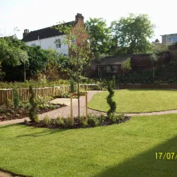 Starannie zaprojektowany ogród z równo przystrzyżonym trawnikiem, ścieżką z kostki brukowej, niskim drewnianym ogrodzeniem i młodym drzewkiem podpartym drewnianą konstrukcją.