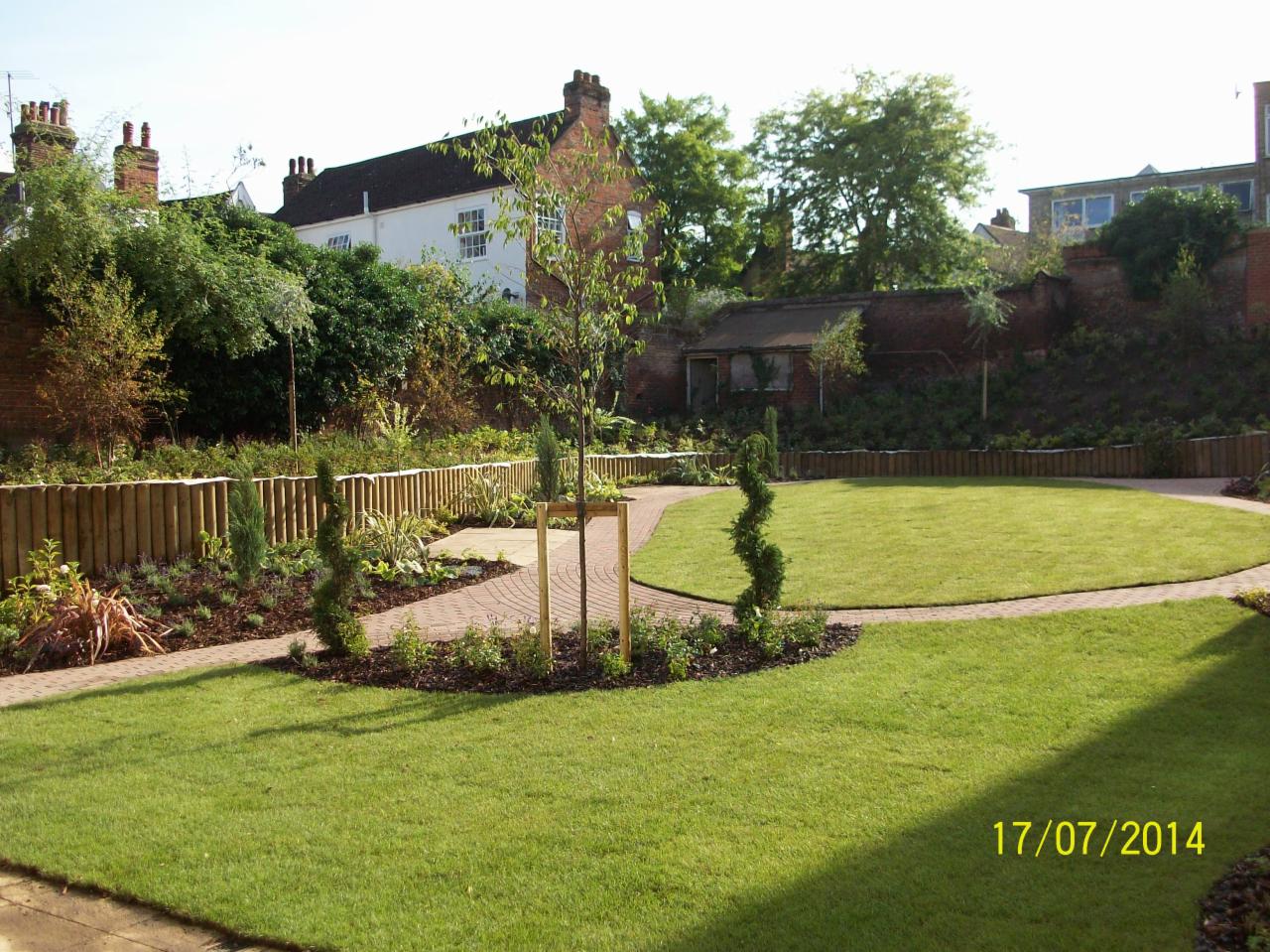 Starannie zaprojektowany ogród z równo przystrzyżonym trawnikiem, ścieżką z kostki brukowej, niskim drewnianym ogrodzeniem i młodym drzewkiem podpartym drewnianą konstrukcją.