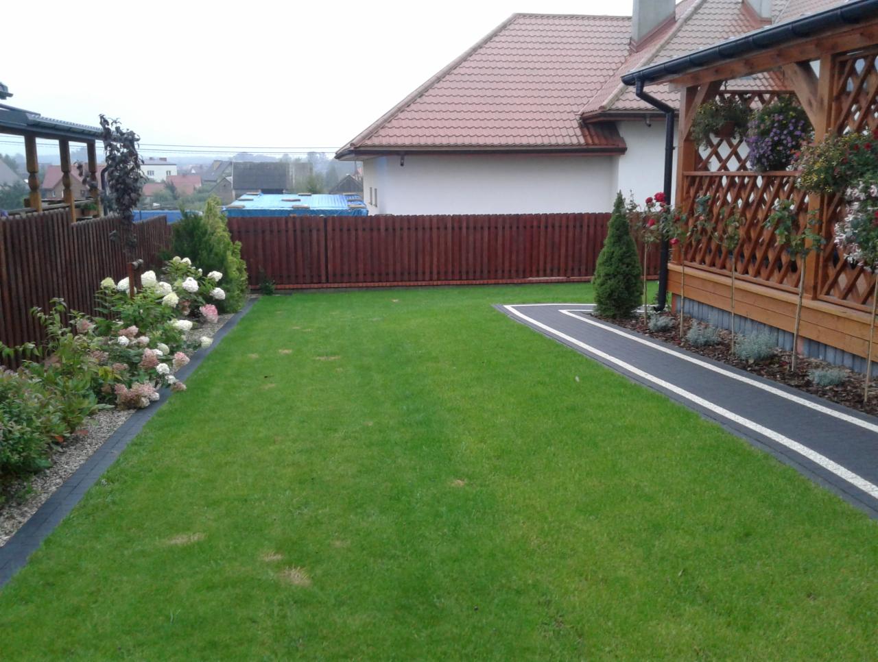 Zadbany trawnik z obrzeżami, rabaty kwiatowe z hortensjami, ścieżka z kostki brukowej prowadząca do drewnianej altany z kratownicą, ogrodzenie drewniane w tle.