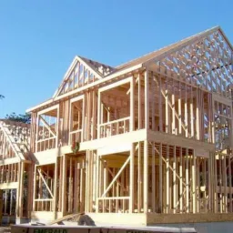 Konstrukcja szkieletowa domu dwukondygnacyjnego z widocznym wieńcem świątecznym na jednym z okien, w tle drzewa i błękitne niebo.