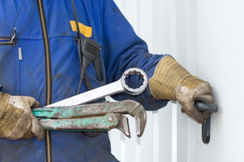 Hydraulik w niebieskim kombinezonie roboczym, z radiem na ramieniu, trzyma dwa klucze: nastawny i płaski, podczas pracy przy rurze na tle białej ściany.