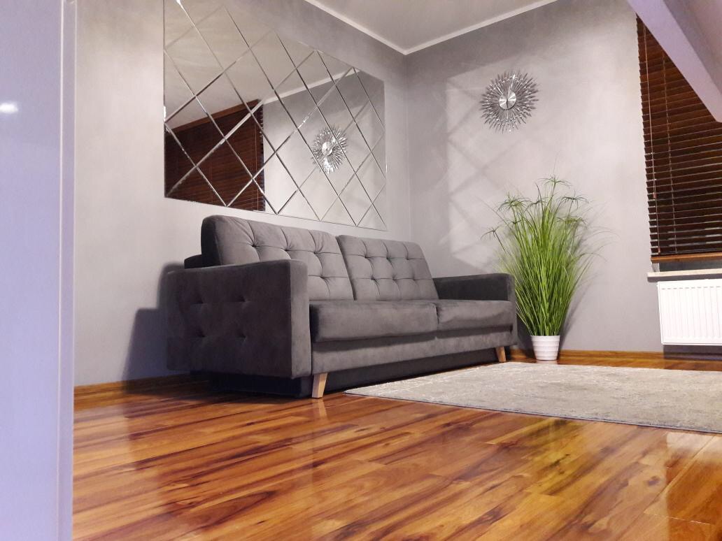 Szara sofa w salonie z lustrzaną mozaiką na ścianie, drewnianą podłogą i rośliną doniczkową, uzupełniona dywanem.