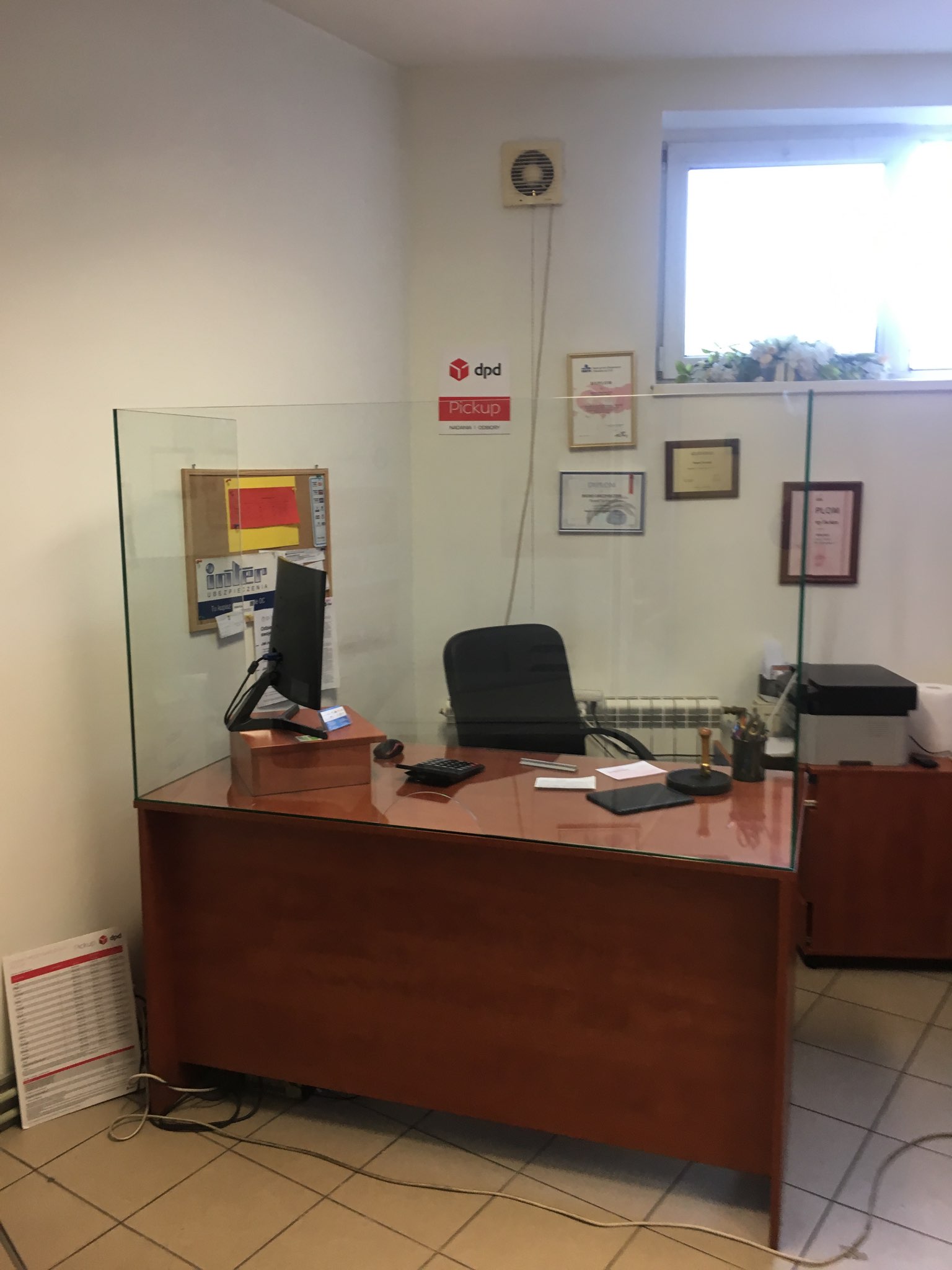 Biuro z biurkiem zabezpieczonym szklaną przegrodą ochronną, monitor, dokumenty i akcesoria biurowe na blacie, ściana z dyplomami i logo firmy kurierskiej w tle.