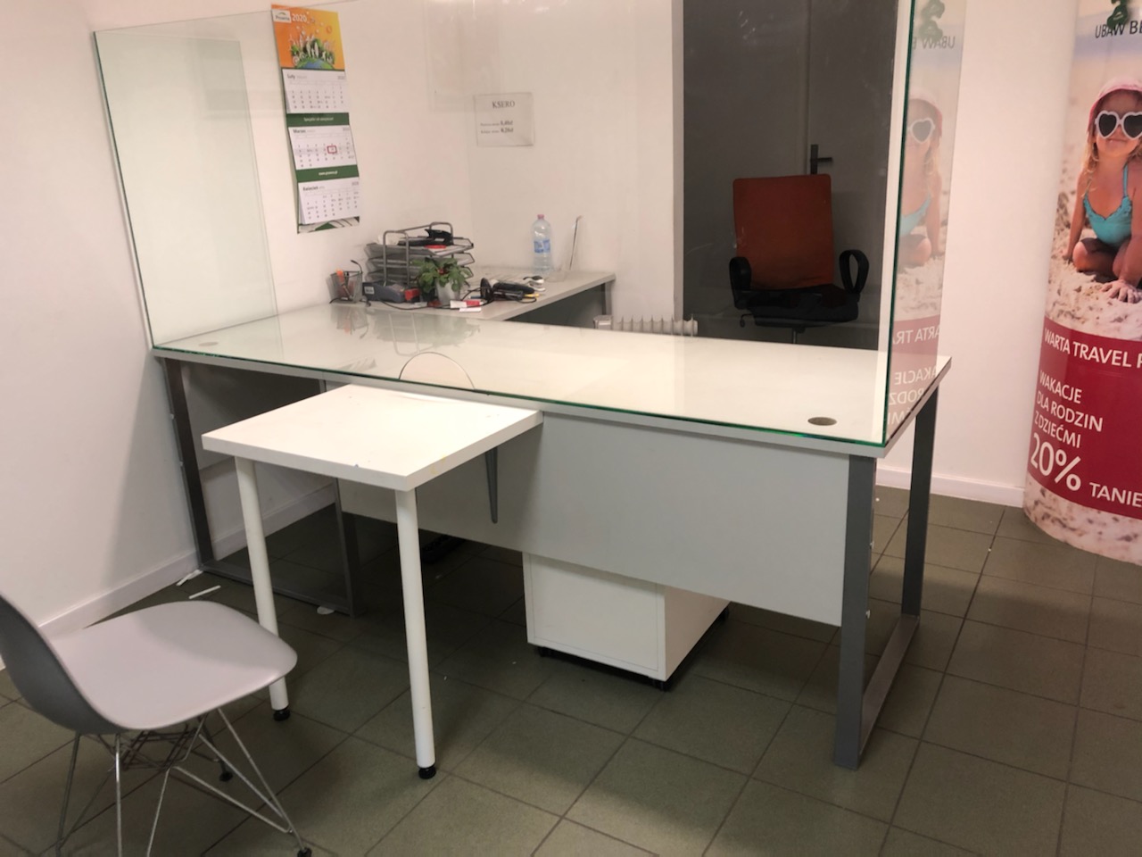 Biuro z przeszklonym biurkiem i przegrodami, widoczny kalendarz i reklama biura podróży