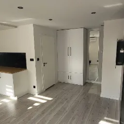 Jasnoszara podłoga z paneli ułożona wzdłuż, z widocznymi promieniami słońca, białe ściany, drzwi i szafa w tle, czarne punkty świetlne w suficie.