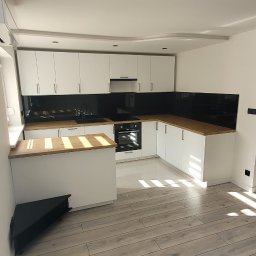 Glazurnictwo-Remonty - Jasna kuchnia z białymi szafkami, drewnianym blatem i czarnym panelem ściennym, oświetlona promieniami słonecznymi padającymi na podłogę z paneli imitujących drewno.