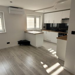 Glazurnictwo-Remonty - Nowoczesna, jasna kuchnia z białymi szafkami, drewnianym blatem i podłogą z paneli w odcieniach szarości, oświetlona promieniami słońca wpadającymi przez okno.