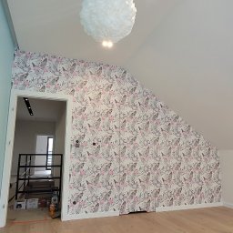 Glazurnictwo-Remonty - Pokój na poddaszu po remoncie, z drewnianą podłogą, ścianą pokrytą tapetą w flamingi i liście oraz wiszącą, białą lampą w kształcie kuli, widoczny fragment klatki schodowej w głębi.