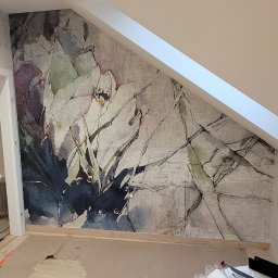 Glazurnictwo-Remonty - Ściana w pokoju na poddaszu pokryta tapetą z motywem kwiatowym w odcieniach szarości, zieleni i fioletu, podłoga zabezpieczona kartonem, widoczne narzędzia i rolka tapety.