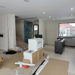 Glazurnictwo-Remonty - Wnętrze w trakcie montażu mebli kuchennych: białe i szare szafki ustawione na kartonach ochronnych, widoczne listwy przypodłogowe, butelka wody i narzędzia. W tle schody, okno i dekoracyjna tapeta.