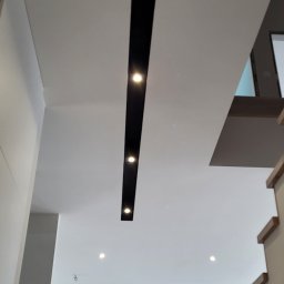 Glazurnictwo-Remonty - Nowoczesne oświetlenie sufitowe z czarną listwą i punktowymi lampami w jasnym wnętrzu z widoczną częścią kuchni i schodami.