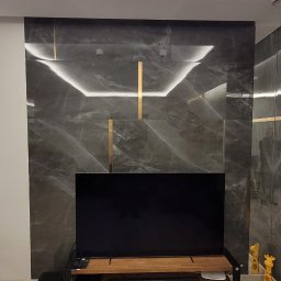 Glazurnictwo-Remonty - Nowoczesny salon z szarą ścianą z efektem marmuru, podświetleniem LED i złotymi paskami, drewnianą podłogą i szafką RTV z czarnymi elementami, widoczne głośniki i dekoracyjne figurki żyraf.