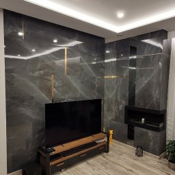 Glazurnictwo-Remonty - Elegancki salon z szarymi płytkami imitującymi marmur na ścianie, podkreślonymi złotymi wstawkami, drewnianą podłogą, telewizorem na szafce RTV i podwieszanym sufitem z oświetleniem LED.