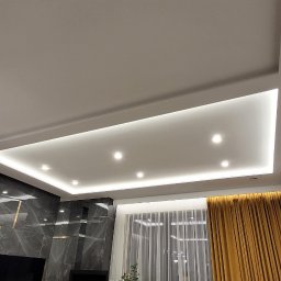 Glazurnictwo-Remonty - Nowoczesny salon z podwieszanym sufitem z oświetleniem LED, szarą ścianą z imitacją marmuru, telewizorem, drewnianą podłogą i szarą sofą z żółtymi poduszkami.