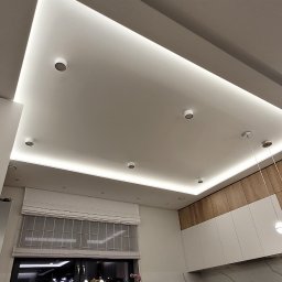 Glazurnictwo-Remonty - Nowoczesna kuchnia z oświetleniem LED w suficie podwieszanym, widoczne okno z roletami rzymskimi, białe szafki i drewniane akcenty.