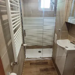 Wąska łazienka po remoncie z kabiną prysznicową z mleczną szybą, grzejnikiem drabinkowym, umywalką z szafką i toaletą, wykończona płytkami w odcieniach brązu i drewnopodobnymi panelami na podłodze.