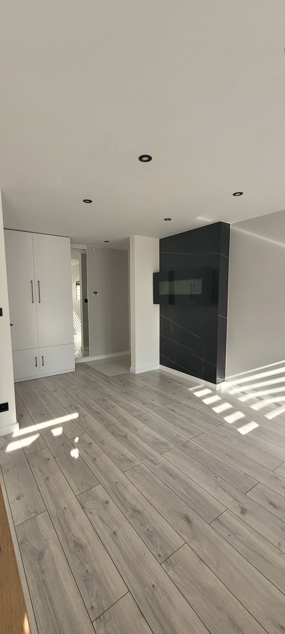Jasnoszara podłoga z paneli laminowanych w przestronnym, minimalistycznym wnętrzu z białą szafą w zabudowie i czarnym panelem dekoracyjnym na ścianie.