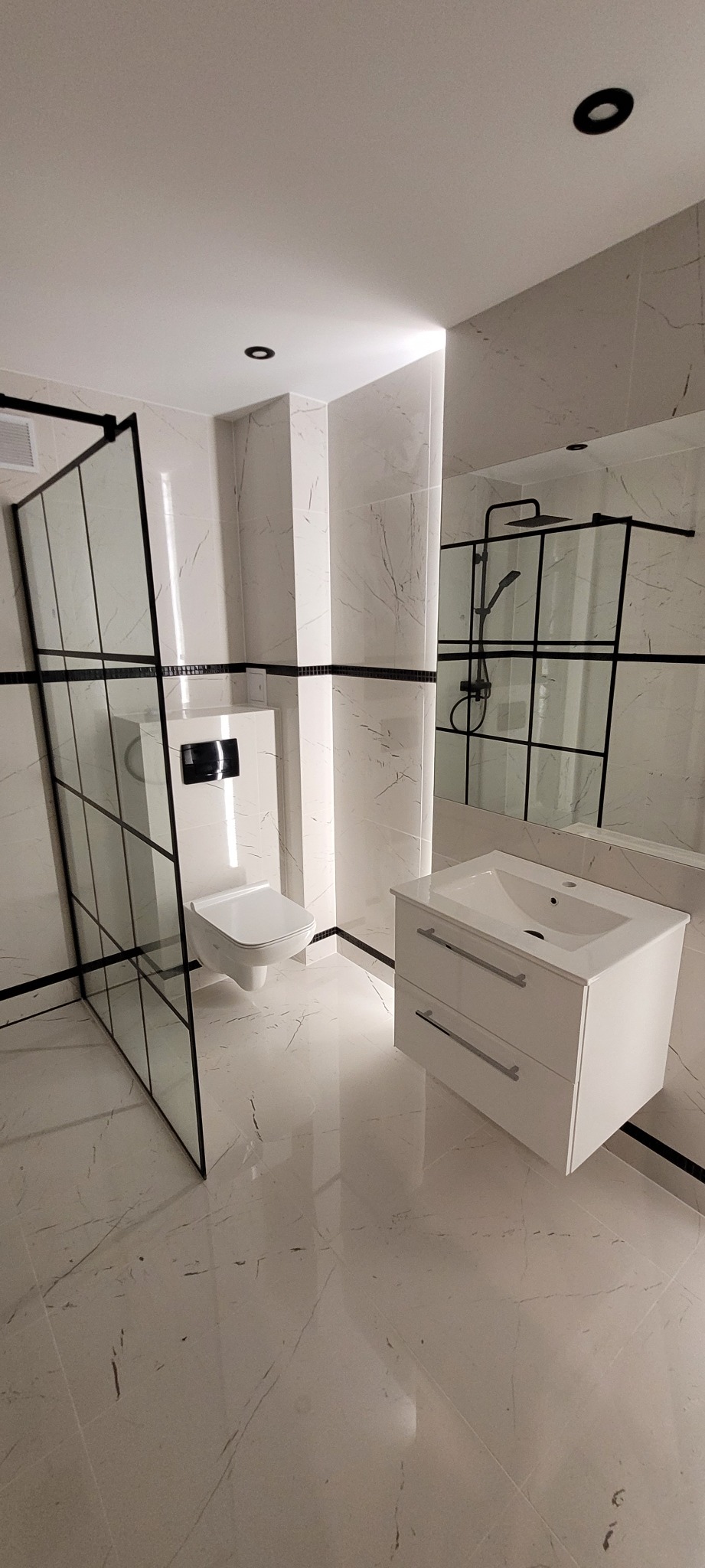 Nowoczesna, minimalistyczna łazienka z białymi marmurowymi płytkami, kabiną prysznicową typu walk-in z czarnymi ramami, podwieszaną toaletą i białą szafką umywalkową z dwiema szufladami...