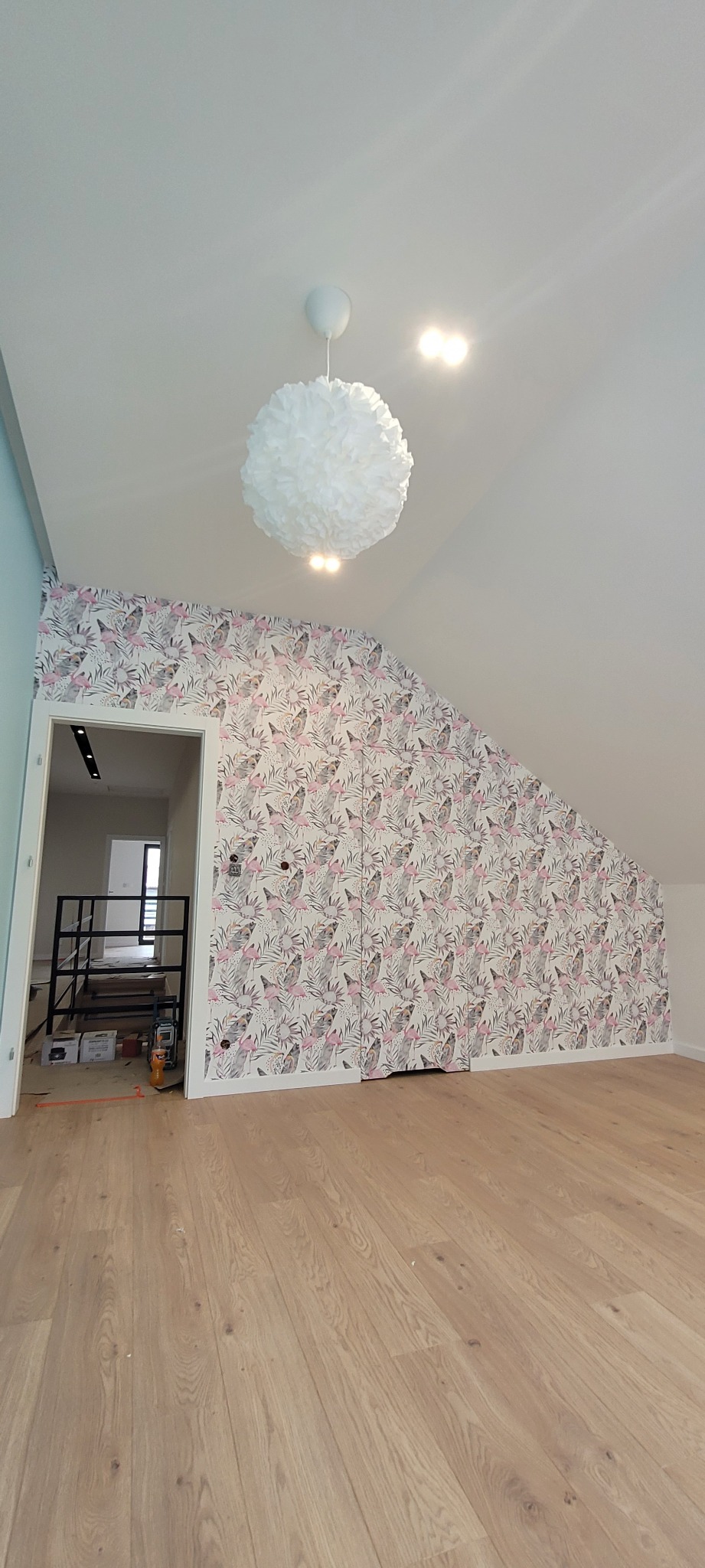 Pokój na poddaszu po remoncie, z drewnianą podłogą, ścianą pokrytą tapetą w flamingi i liście oraz wiszącą, białą lampą w kształcie kuli, widoczny fragment klatki schodowej w głębi.