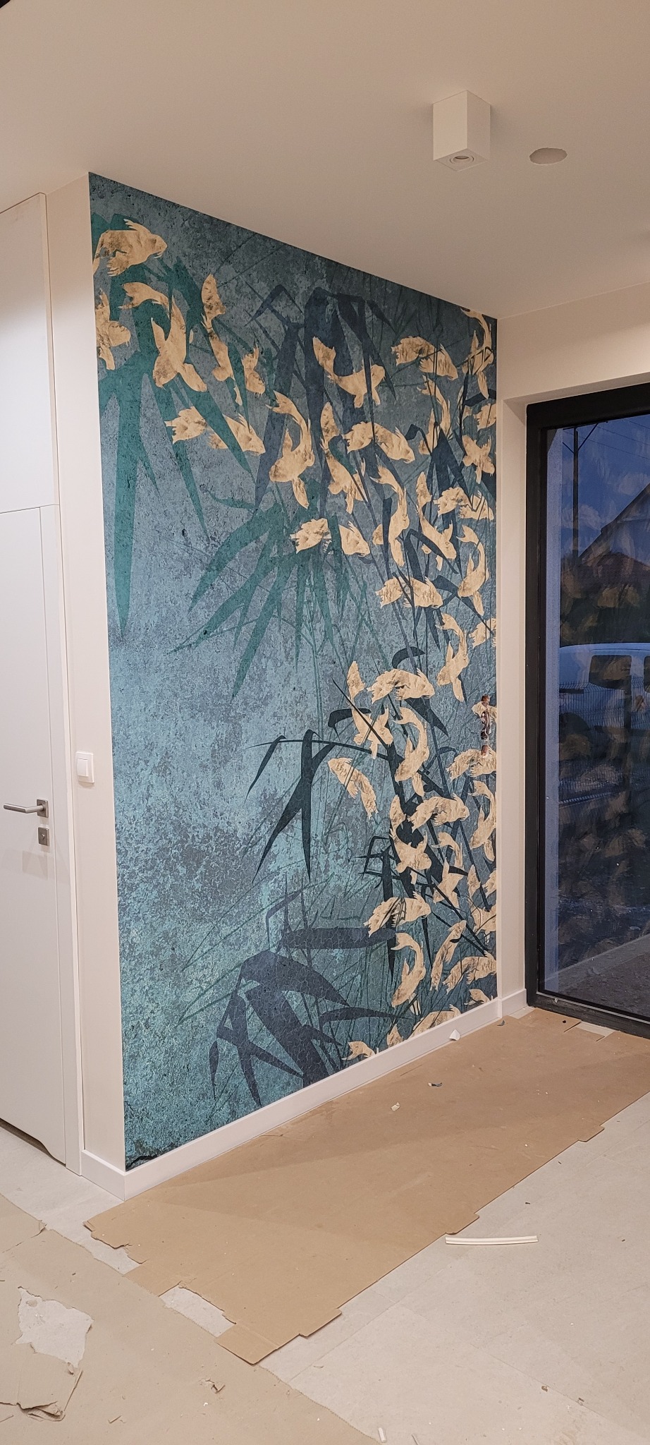Ściana z tapetą w odcieniach turkusu i złota z motywem bambusa i liści, widok na fragment podłogi zabezpieczonej kartonem podczas prac wykończeniowych.